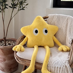 May include: A bright yellow plush star-shaped cushion with a soft, fuzzy texture. The cushion features two large white oval eyes with black pupils, two long arms with three-fingered hands, and two long legs extending downwards from the main body.