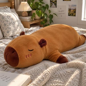 May include: A plush capybara toy with soft brown fur, a dark brown snout, and small ears. The stuffed animal is lying in a prone position with its eyes closed, featuring embroidered facial details and dark brown accents on its paws.