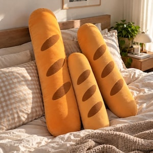 May include: Three plush pillows shaped like French baguettes in varying sizes. Each pillow is golden yellow with dark brown oval-shaped accents along the top, mimicking the appearance of scored bread crusts on a soft, textured fabric surface.