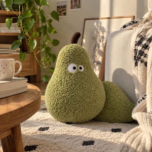 May include: A pear-shaped plush toy with a textured olive green exterior, a dark brown stem, and two white circular eyes with black pupils. The plush has a soft, fuzzy surface and a rounded base, sitting upright with a smaller matching green section.