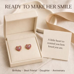 Könnte beinhalten: Ein Paar herzförmige Ohrstecker mit rosa und weißen Steinen in einer beigen Schmuckschatulle. Eine Karte zeigt den Text: A little heart to remind you how loved you are. Zudem sind die Texte READY TO MAKE HER SMILE sowie Birthday, Best Friend, Daughter, Anniversary zu sehen.