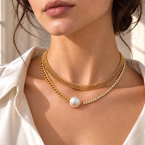 Könnte beinhalten: Eine mehrlagige goldfarbene Halskette bestehend aus einer mehrreihigen Perlenkette und einer zweiten Kette mit einem goldfarbenen Gliederelement, einer einzelnen großen weißen Perle und einer Reihe klarer Kristalle auf einer Metallbasis.