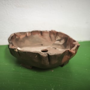 Puede incluir: Una maceta para bonsái de cerámica poco profunda y moldeada a mano con un esmalte moteado de color marrón oscuro. El borde presenta una forma irregular y ondulada, y la base incluye pequeñas patas y un orificio de drenaje central rústico.