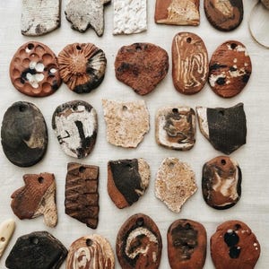 Puede incluir: Una colección de veinticinco colgantes de arcilla planos e irregulares dispuestos en cinco filas. Cada pieza presenta un orificio único para colgar y muestra texturas variadas, tonos tierra de marrón, negro, blanco y beige, y patrones únicos.