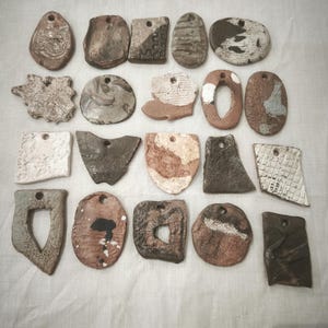Może przedstawiać: Kolekcja dwudziestu różnorodnych ceramicznych zawieszek o różnych kształtach, w tym koła, prostokąty i formy nieregularne. Każdy element posiada unikalne tekstury, ziemiste odcienie brązu, szarości i bieli oraz otwór do zawieszenia.