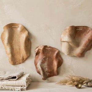 Puede incluir: Tres placas de pared de formas orgánicas irregulares en tonos beige, marrón claro y terracota. Cada pieza presenta una superficie texturizada y un pequeño lazo horizontal en la parte superior para colgar, junto a textiles beige doblados.