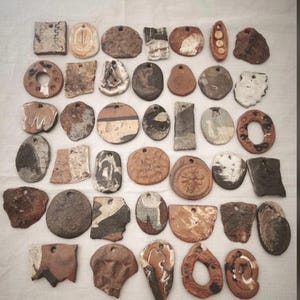 Puede incluir: Una colección de treinta y cinco colgantes de cerámica variados en diversas formas, incluyendo círculos, cuadrados y formas irregulares. Estas piezas de arcilla presentan texturas diversas, tonos tierra marrones y grises, y algunos incluyen motivos de abejas.