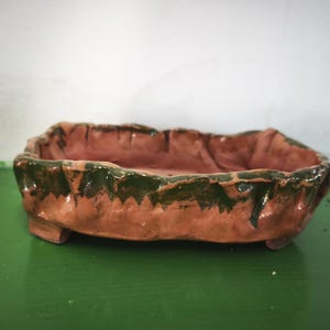 Puede incluir: Una maceta rectangular de cerámica para bonsái con un borde irregular formado a mano. El exterior y el interior presentan una base de arcilla color terracota con un esmalte verde oscuro que gotea desde el borde superior por los lados.