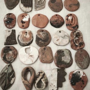 Può includere: Una collezione di venticinque ciondoli in ceramica fatti a mano disposti in cinque file. Ogni ciondolo presenta forme organiche uniche, texture variegate e palette di colori terrosi che includono tonalità marroni, bianche, nere e grigie, con fori.