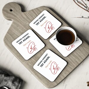 May include: Four square white coasters featuring the black text WRONG ANSWER? TRY AGAIN. and That's maths. in a smaller font. Each coaster displays a red circled checkmark graphic. One coaster supports a white ceramic mug filled with dark coffee.