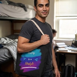 Op de afbeelding: Een zwarte tote bag met een kleurrijk penseelstreekontwerp in tinten blauw, paars en magenta. De tekst "Writing is a lifestyle" is in wit en goudkleurig cursief gedrukt in het midden van de afbeelding op de voorzijde van de tas.