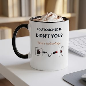 May include: A white ceramic mug with a black handle and rim, filled with whipped cream. The front features the black text "YOU TOUCHED IT. DIDN'T YOU?" above "That's technology." and a graphic of a dial, a cable, and a port connector.