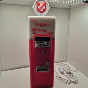 Könnte beinhalten: Ein rot-weißer Jugger-Nog-Verkaufsautomaten-Nachbau mit einem runden Schild oben mit rotem Kreuz, dem Text Drink Jugger-Nog, That's a good nog, 10 cents, Made with real eggs, einem Münzschlitz und einem weißen USB-Stromkabel.