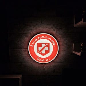 Könnte beinhalten: Ein kreisförmiges Wandleuchtschild in Rot und Weiß. Das Schild zeigt den Schriftzug Juggernog Soda in einer stilisierten Schriftart, der ein zentrales rotes Schildelement mit einem weißen Kreuz und einer diagonalen Patronengrafik umgibt.