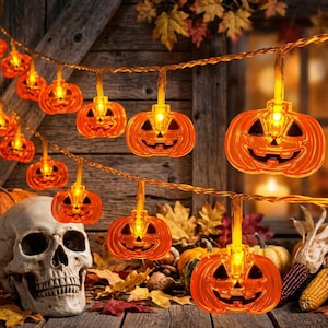 May include: A string of orange jack-o'-lantern pumpkin lights with glowing internal bulbs. The translucent plastic pumpkins feature carved facial expressions and are connected by a thin wire. A human skull model sits nearby.