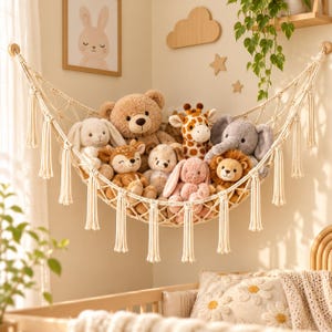 May include: A cream-colored macrame toy hammock with decorative tassels hanging on a wall, filled with a collection of plush stuffed animals including a brown bear, a giraffe, an elephant, a lion, and several small bunnies and a deer.