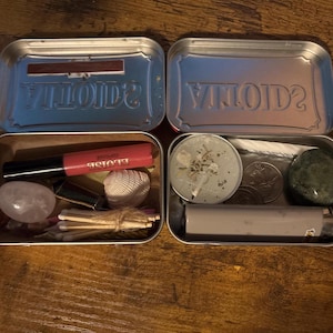 Op de afbeelding: Twee open metalen Altoids-blikjes met diverse voorwerpen. Het linkerblikje bevat een roze lipgloss, een witte schelp, een gladde roze steen en een bundeltje lucifers. Het rechterblikje bevat een kleine ronde kaars, een munt en een groene steen.