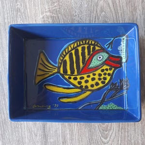 Op de afbeelding: Een rechthoekig blauw keramieken dienblad met een handgeschilderde gele vis met zwarte strepen en stippen, een rode mond en een groen oog. Het ontwerp bevat zwarte waterplanten en de handtekening Corneille '99 in de linkerbenedenhoek.