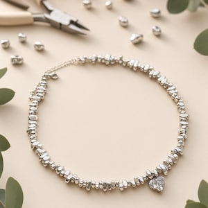 Puede incluir: Un collar de tono plateado compuesto por pequeñas cuentas metálicas irregulares ensartadas, con un único colgante en forma de corazón incrustado con piedras transparentes. El collar incluye una fina cadena metálica con cierre.
