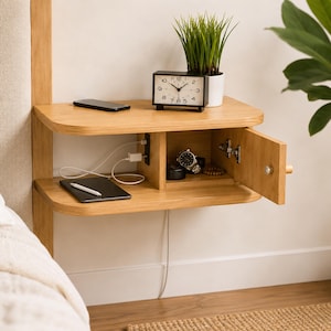 May include: A light oak floating nightstand with two shelves, a built-in power outlet, and a small cabinet door. The top shelf holds a black analog clock and a potted plant. The lower shelf holds a black tablet with a stylus and a charging cable.