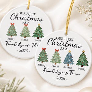 Può includere: Due ornamenti rotondi in ceramica bianca con cordini dorati. Ognuno presenta alberi di Natale ad acquerello, il testo "OUR FIRST Christmas AS A", "Family of Three" o "Family of Four", nomi e l'anno "2026" stampati in inchiostro nero.