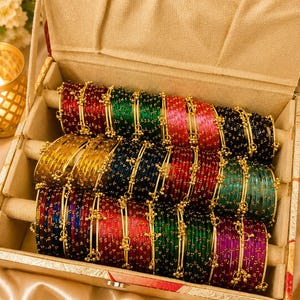 Puede incluir: Una colección de brazaletes de vidrio multicolor dispuestos en un joyero forrado de tela beige. Los brazaletes presentan un acabado translúcido en tonos rojo, verde, azul, amarillo y morado, acentuados con pequeñas cuentas de metal dorado.