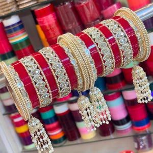 May include: A set of red and gold bangles featuring alternating bands of solid red, rows of clear stones in gold settings, and thin gold beaded strands. Three gold bell-shaped charms with white beads hang from the bangles on a metal rod.