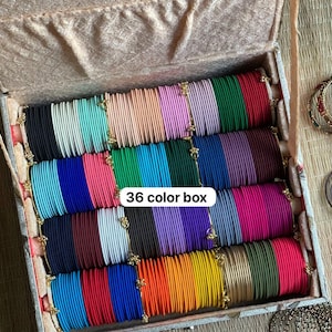 Könnte beinhalten: Eine rechteckige Aufbewahrungsbox mit einer Sammlung dünner Metallarmreifen, die in Reihen nach Farben angeordnet sind. Das Set enthält 36 Farbvarianten in den Tönen Schwarz, Blau, Pink, Grün, Rot, Lila, Orange, Gelb und metallischem Gold.