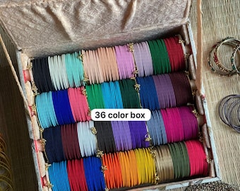 May include: A rectangular storage box containing a collection of thin metal bangles arranged in rows by color. The set includes 36 color box variations in shades of black, blue, pink, green, red, purple, orange, yellow, and metallic gold.