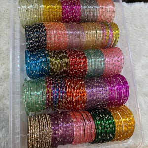 Pode incluir: Uma coleção de pulseiras circulares finas dispostas em filas dentro de um recipiente de plástico transparente. Cada pulseira apresenta um acabamento translúcido ou metálico em várias cores, incluindo ouro, rosa, azul, verde e preto, acentuado com pequenos tachas prateadas espaçadas uniformemente.
