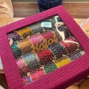 May include: A square, dark pink textured gift box with a clear window lid containing multiple rows of thin, metallic bangles in various colours including red, green, gold, and blue. The name Kajol is printed in gold script on the clear window lid.