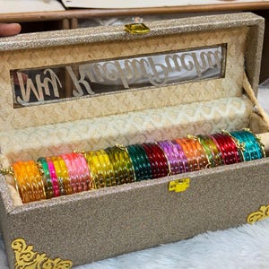 May include: A rectangular, glittery gold storage box with gold corner accents and a clear window on the lid featuring white script text. Inside, a row of translucent, multi-coloured glass bangles in shades of orange, pink, yellow, green, and red.