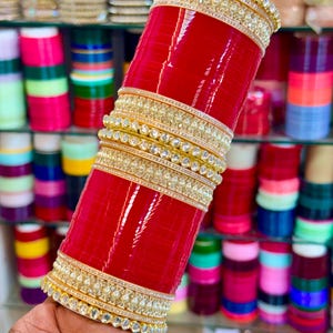 Peut inclure: Un ensemble de bracelets rouges avec des bandes dorées incrustées de pierres transparentes. La pile comprend plusieurs bracelets rouges unis alternés avec des bandes décoratives dorées, formant un cylindre à la finition polie et brillante.
