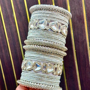 Pode incluir: Uma pilha de pulseiras prateadas com um acabamento texturizado e brilhante. O conjunto inclui vários aros finos e uniformes intercalados com duas faixas mais largas decoradas com um padrão repetitivo de pedras triangulares transparentes.