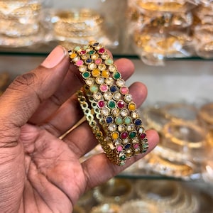 Peut inclure: Un ensemble de trois bracelets joncs en métal doré présentant un motif répétitif de petites pierres cabochons rondes et multicolores dans des tons rose, jaune, vert et bleu, alternées avec des cristaux circulaires transparents et facettés.
