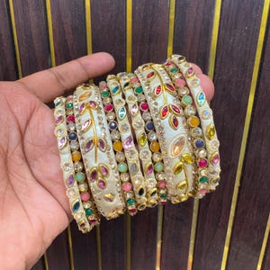 Op de afbeelding: Een set crèmekleurige armbanden met goudkleurige metalen bladmotieven en ingelegde veelkleurige strass-steentjes in rood, blauw, groen, roze en geel. De armbanden zijn gestapeld en tonen gedetailleerd metaalwerk en diverse gekleurde steentjes.