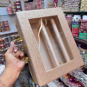 May include: A rectangular, glittery gold jewelry display case featuring a clear glass lid and two internal velvet-lined ring rolls. The box includes a gold-toned metal handle on the side for carrying and a secure latch mechanism for closure.