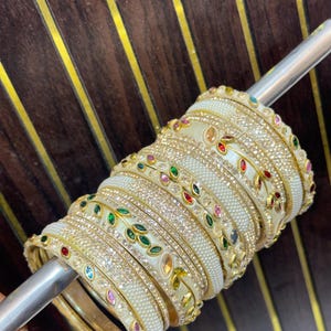 May include: A set of stacked bangles featuring a cream-colored base with embedded white pearls and clear crystals. The bracelets are decorated with gold-tone metal leaf motifs inlaid with small, multi-colored gemstones in red, green, and blue.