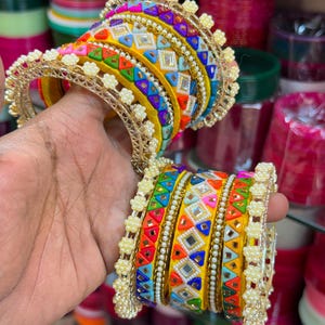 May include: A set of bangles featuring yellow fabric bands decorated with colorful geometric shapes, small mirrors, and white beaded edges. The bracelets are arranged in two groups, showcasing a vibrant pattern of red, blue, green, and orange accents.