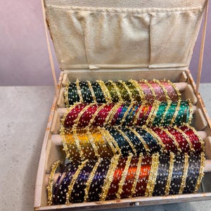 Op de afbeelding: Een verzameling veelkleurige metalen armbanden gerangschikt in een met beige stof beklede juwelendoos. Elke armband is voorzien van een dunne goudkleurige rand met kleine, verhoogde decoratieve accenten. De set bevat diverse kleuren zoals groen, geel, rood, blauw, paars en roze, georganiseerd in rijen op gevoerde horizontale stangen.