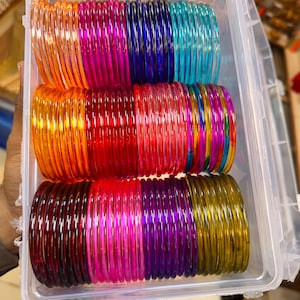 May include: A clear plastic rectangular storage container filled with rows of thin, translucent, circular bangles in various vibrant colours including orange, magenta, deep blue, turquoise, red, purple, and yellow, arranged in neat stacks.