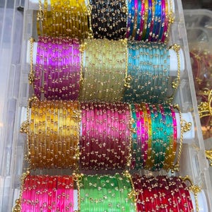 Peut inclure: Une collection de bracelets joncs fins en verre translucide disposés dans une boîte de rangement en plastique transparent. Les bracelets présentent un motif répétitif de petites fleurs dorées métalliques et sont disponibles dans une variété de couleurs vives, notamment jaune, noir, bleu, rose, vert et rouge.