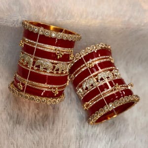 May include: Two wide red bangles featuring gold-tone metal bands, clear rhinestone embellishments, and decorative elephant motifs. The bangles are accented with small dangling gold charms and rows of sparkling crystals along the top and bottom edges.