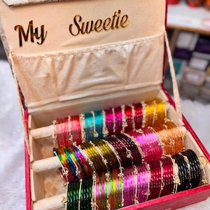 May include: A red jewellery box with a floral lid and the text My Sweetie, containing three horizontal velvet-covered rods holding rows of thin, translucent, multi-coloured bangles in shades of red, blue, pink, yellow, green, and black.
