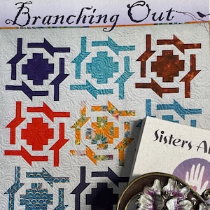 May include: A quilt pattern titled "Branching Out" with a colorful geometric design. The pattern features blocks in shades of purple, orange, teal, and brown. A decorative floral brooch and a business card from "Sisters Artisans" are also in the image.