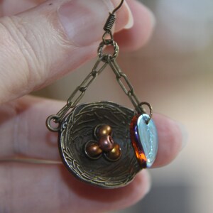 May include: A bronze earring with a small bird's nest charm, three copper beads, and a blue and purple iridescent glass teardrop charm.