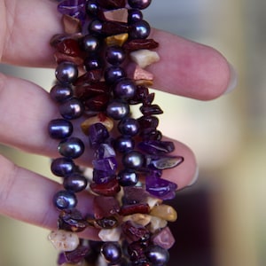May include: A multi-strand necklace with dark purple pearls and various shades of purple, red, and brown stones.