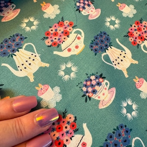 May include: Teal fabric with a repeating pattern of white teapots filled with colorful flowers. The teapots have a gold star design and are surrounded by white stars.
