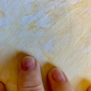 May include: Close-up of two fingers on a yellow fabric with a white and yellow tie-dye pattern.