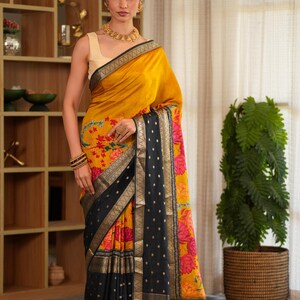 May include: A mustard yellow and black saree featuring a floral pattern with pink and orange flowers. The garment includes a wide black border with gold metallic detailing and small gold dots, paired with a sleeveless cream-colored blouse.